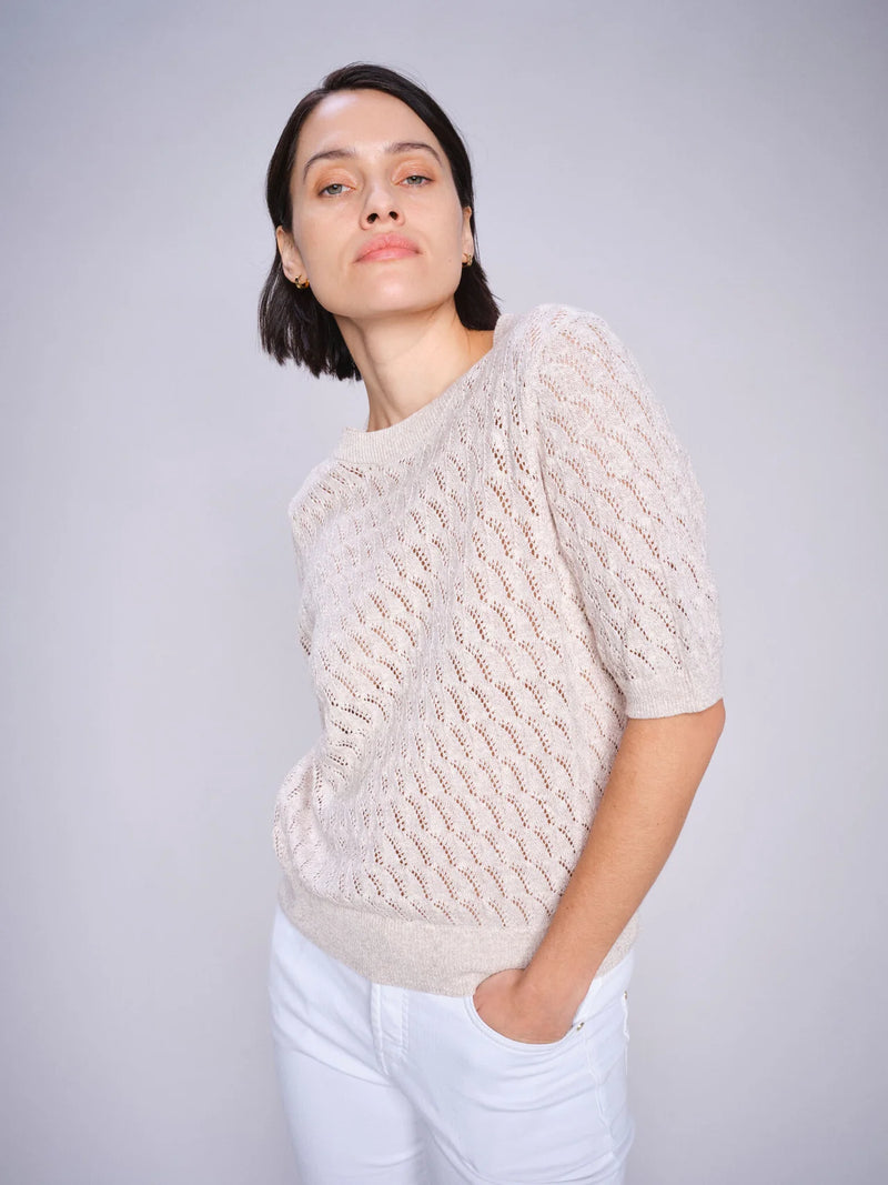 A women's short-sleeve knit sweater in a soft, neutral beige color called Feather Gray. The fabric is lightweight and delicate, featuring an intricate open-knit pattern with a repeating wavy design, allowing for breathability and a slightly sheer effect. The sweater has a classic round neckline with a finely ribbed collar that blends seamlessly into the knit. The short sleeves fall just above the elbow, offering a relaxed and comfortable fit. The hem is finished with a slightly thicker ribbed band, giving t