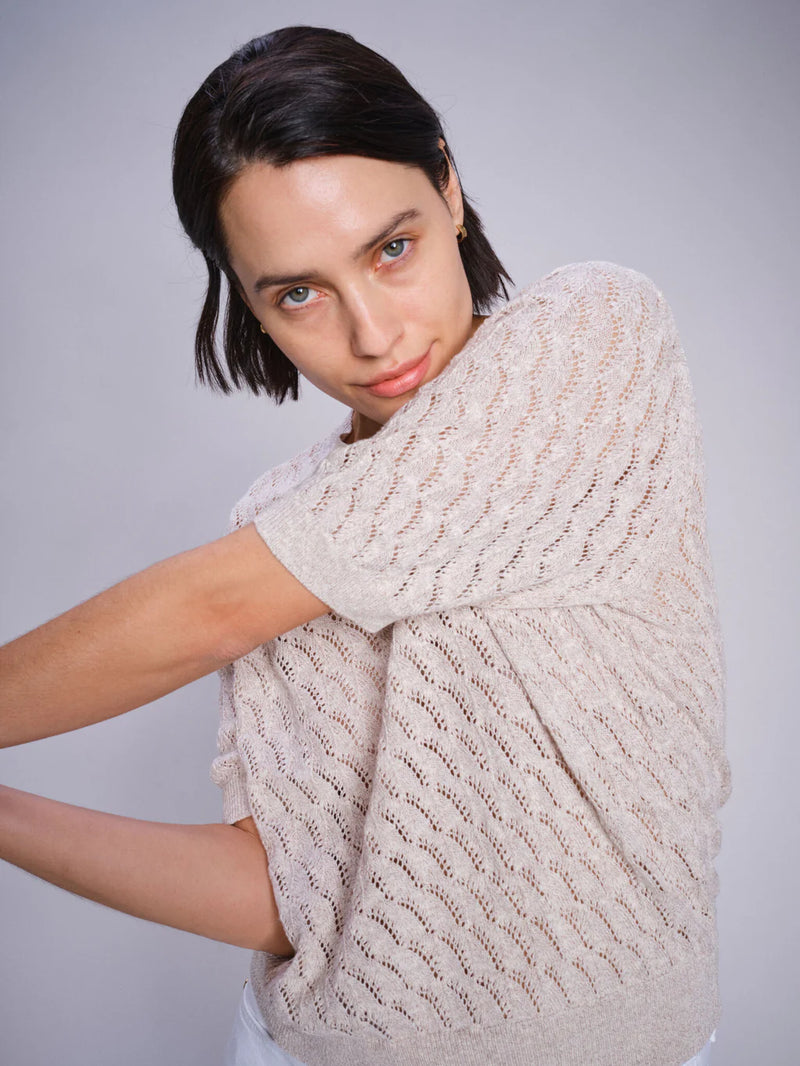 A women's short-sleeve knit sweater in a soft, neutral beige color called Feather Gray. The fabric is lightweight and delicate, featuring an intricate open-knit pattern with a repeating wavy design, allowing for breathability and a slightly sheer effect. The sweater has a classic round neckline with a finely ribbed collar that blends seamlessly into the knit. The short sleeves fall just above the elbow, offering a relaxed and comfortable fit. The hem is finished with a slightly thicker ribbed band, giving t