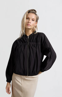 A women's lightweight bomber jacket in a soft beige color with a relaxed, slightly oversized fit. It features a round neckline, a full-length front zipper, and gathered detailing along the chest for a subtle textured effect. The long sleeves are voluminous and taper into elasticated cuffs, while the hem also has an elastic finish for a comfortable and slightly cropped silhouette.