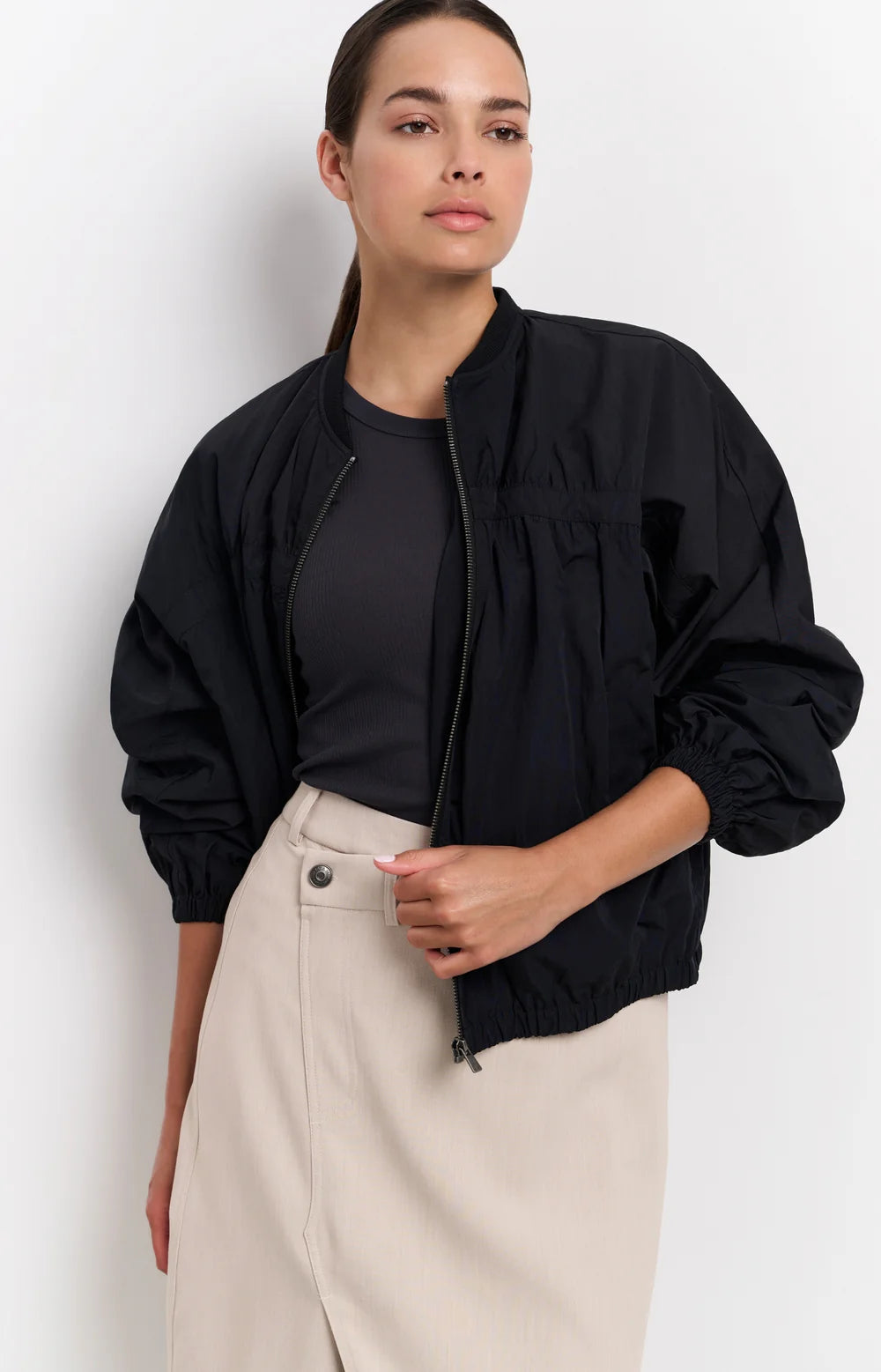 A women's lightweight bomber jacket in a soft beige color with a relaxed, slightly oversized fit. It features a round neckline, a full-length front zipper, and gathered detailing along the chest for a subtle textured effect. The long sleeves are voluminous and taper into elasticated cuffs, while the hem also has an elastic finish for a comfortable and slightly cropped silhouette.
