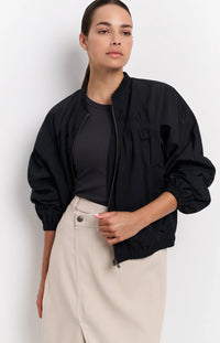 A women's lightweight bomber jacket in a soft beige color with a relaxed, slightly oversized fit. It features a round neckline, a full-length front zipper, and gathered detailing along the chest for a subtle textured effect. The long sleeves are voluminous and taper into elasticated cuffs, while the hem also has an elastic finish for a comfortable and slightly cropped silhouette.