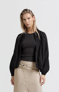 A women's lightweight bomber jacket in a soft beige color with a relaxed, slightly oversized fit. It features a round neckline, a full-length front zipper, and gathered detailing along the chest for a subtle textured effect. The long sleeves are voluminous and taper into elasticated cuffs, while the hem also has an elastic finish for a comfortable and slightly cropped silhouette.