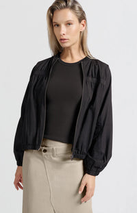 A women's lightweight bomber jacket in a soft beige color with a relaxed, slightly oversized fit. It features a round neckline, a full-length front zipper, and gathered detailing along the chest for a subtle textured effect. The long sleeves are voluminous and taper into elasticated cuffs, while the hem also has an elastic finish for a comfortable and slightly cropped silhouette.