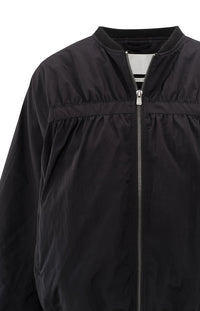 A women's lightweight bomber jacket in a soft beige color with a relaxed, slightly oversized fit. It features a round neckline, a full-length front zipper, and gathered detailing along the chest for a subtle textured effect. The long sleeves are voluminous and taper into elasticated cuffs, while the hem also has an elastic finish for a comfortable and slightly cropped silhouette.