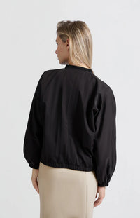 A women's lightweight bomber jacket in a soft beige color with a relaxed, slightly oversized fit. It features a round neckline, a full-length front zipper, and gathered detailing along the chest for a subtle textured effect. The long sleeves are voluminous and taper into elasticated cuffs, while the hem also has an elastic finish for a comfortable and slightly cropped silhouette.