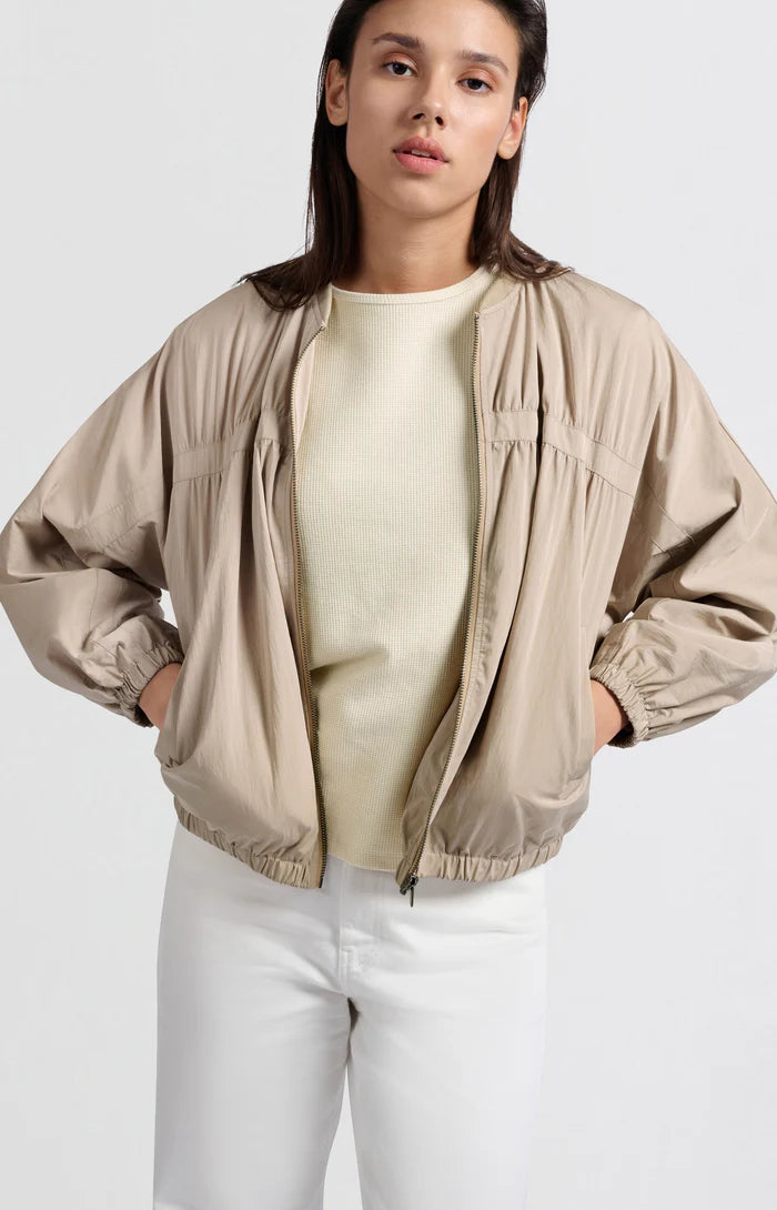 A women's lightweight bomber jacket in a soft beige color with a relaxed, slightly oversized fit. It features a round neckline, a full-length front zipper, and gathered detailing along the chest for a subtle textured effect. The long sleeves are voluminous and taper into elasticated cuffs, while the hem also has an elastic finish for a comfortable and slightly cropped silhouette.