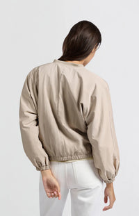A women's lightweight bomber jacket in a soft beige color with a relaxed, slightly oversized fit. It features a round neckline, a full-length front zipper, and gathered detailing along the chest for a subtle textured effect. The long sleeves are voluminous and taper into elasticated cuffs, while the hem also has an elastic finish for a comfortable and slightly cropped silhouette.