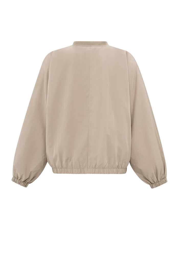 A women's lightweight bomber jacket in a soft beige color with a relaxed, slightly oversized fit. It features a round neckline, a full-length front zipper, and gathered detailing along the chest for a subtle textured effect. The long sleeves are voluminous and taper into elasticated cuffs, while the hem also has an elastic finish for a comfortable and slightly cropped silhouette.