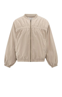 A women's lightweight bomber jacket in a soft beige color with a relaxed, slightly oversized fit. It features a round neckline, a full-length front zipper, and gathered detailing along the chest for a subtle textured effect. The long sleeves are voluminous and taper into elasticated cuffs, while the hem also has an elastic finish for a comfortable and slightly cropped silhouette.