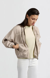 A women's lightweight bomber jacket in a soft beige color with a relaxed, slightly oversized fit. It features a round neckline, a full-length front zipper, and gathered detailing along the chest for a subtle textured effect. The long sleeves are voluminous and taper into elasticated cuffs, while the hem also has an elastic finish for a comfortable and slightly cropped silhouette.