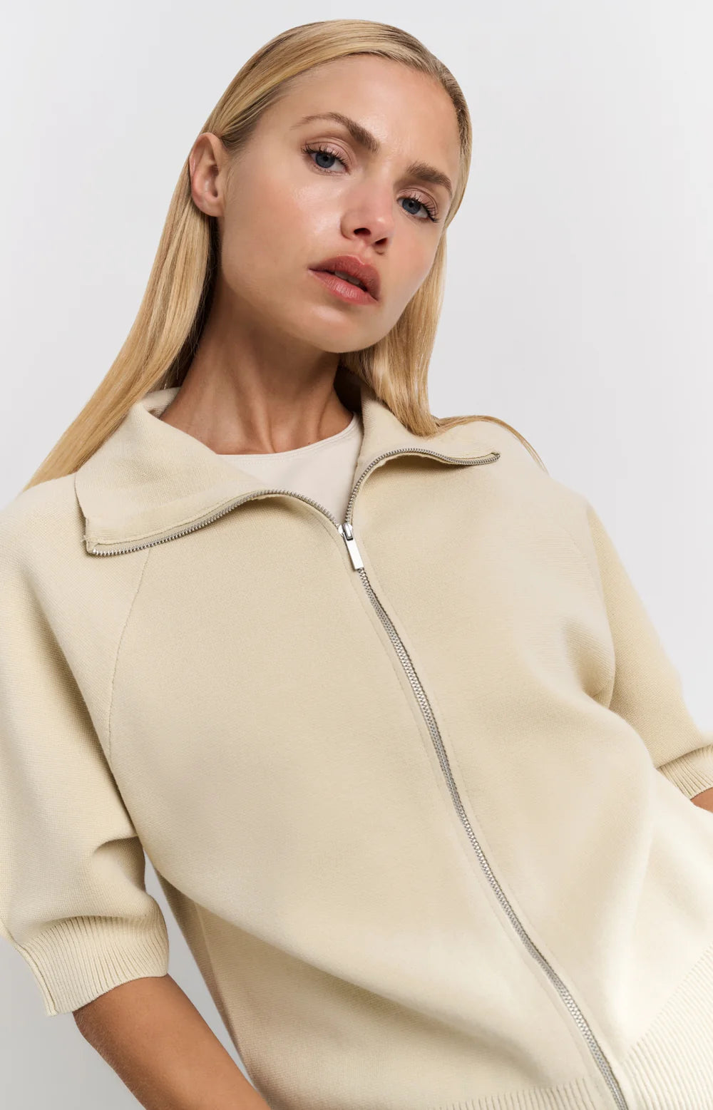 his is a women's short-sleeve zip-up cardigan in a soft, warm beige color. It is made from a finely knitted fabric with a smooth texture and a structured yet comfortable fit. The cardigan features a classic folded collar that sits neatly around the neckline, leading down to a silver-toned full-length zipper at the front, allowing it to be worn open or closed.

The sleeves are elbow-length with a slightly loose fit, finished with ribbed cuffs for a subtle contrast in texture. The hem is also ribbed, giving t