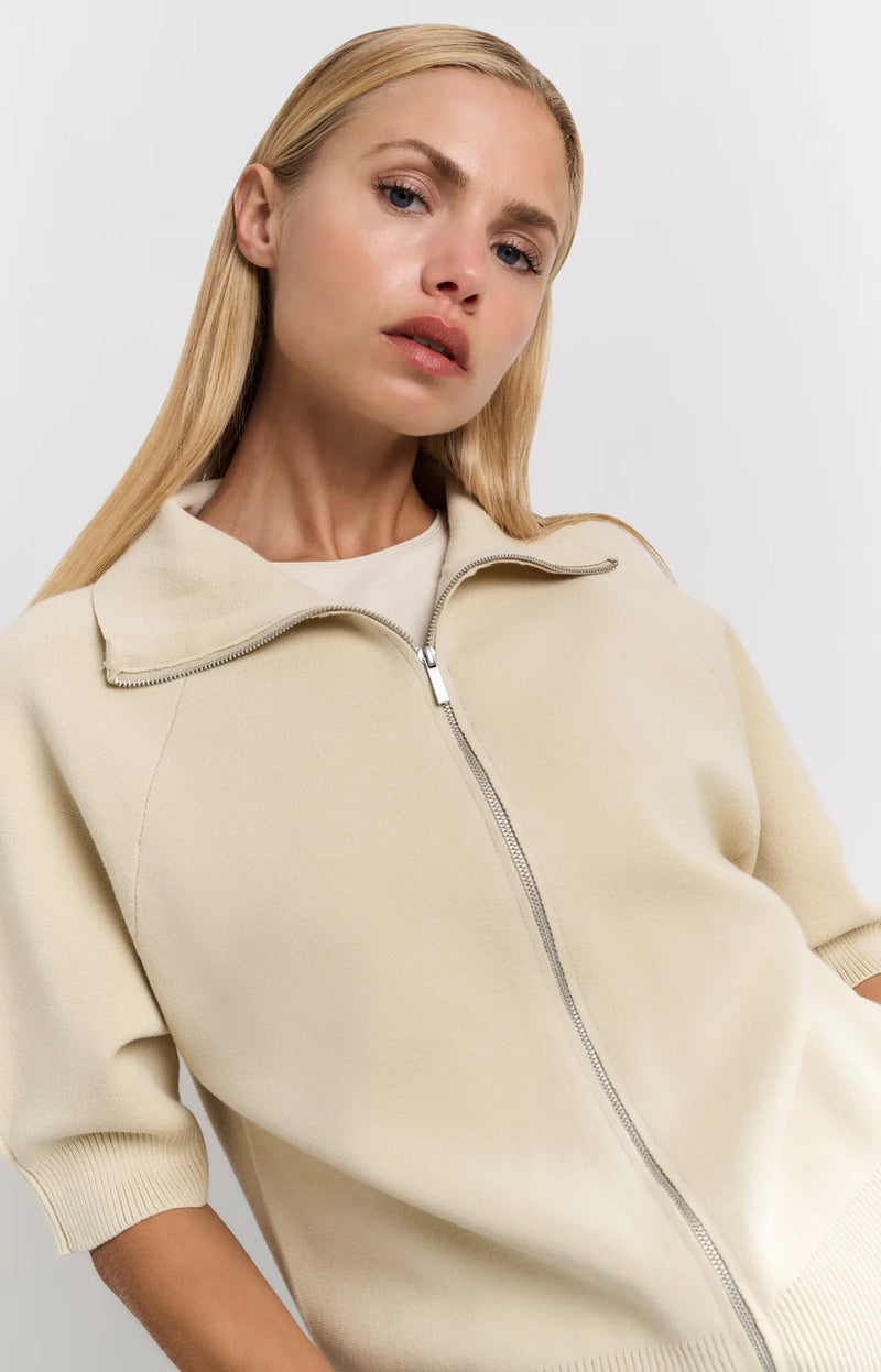 his is a women's short-sleeve zip-up cardigan in a soft, warm beige color. It is made from a finely knitted fabric with a smooth texture and a structured yet comfortable fit. The cardigan features a classic folded collar that sits neatly around the neckline, leading down to a silver-toned full-length zipper at the front, allowing it to be worn open or closed.

The sleeves are elbow-length with a slightly loose fit, finished with ribbed cuffs for a subtle contrast in texture. The hem is also ribbed, giving t